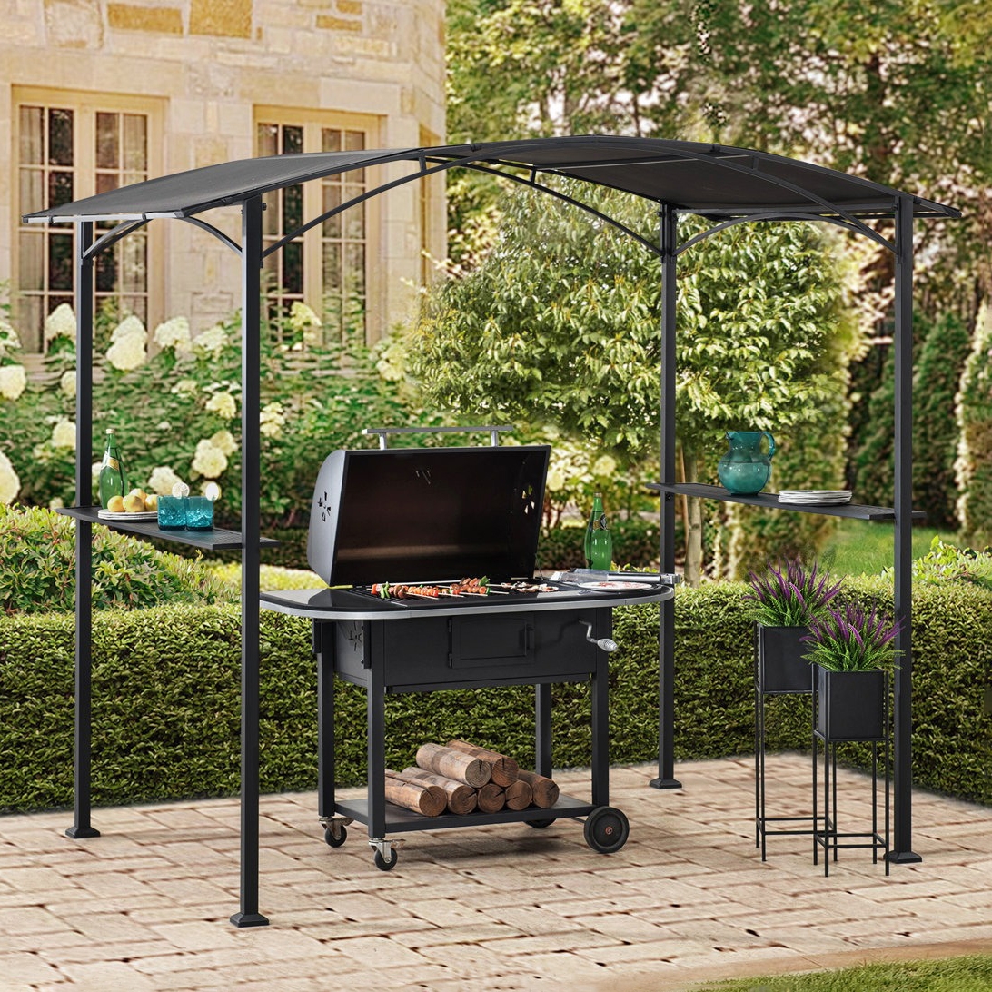 A black metal gazebo with a curved roof shades a charcoal grill, featuring shelves with drinks and dishes, set on a paved patio with logs underneath.