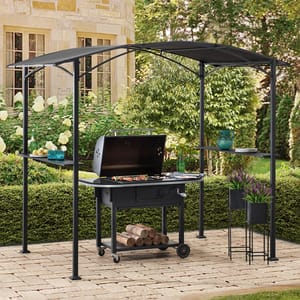 A black metal gazebo with a curved roof shades a charcoal grill, featuring shelves with drinks and dishes, set on a paved patio with logs underneath.
