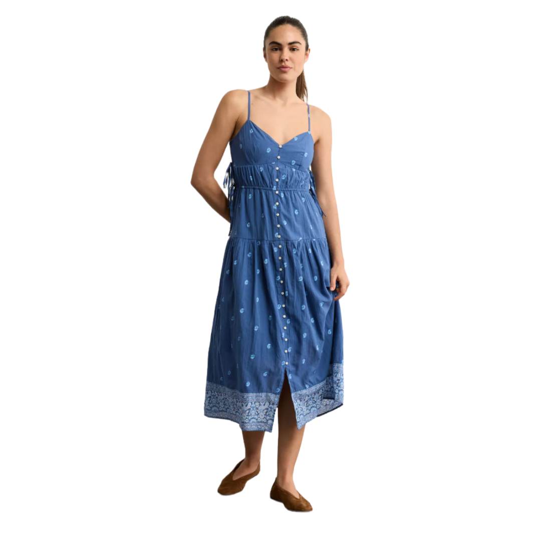 A woman is wearing a sleeveless, blue button-up sundress with white floral patterns and a detailed hem. She's also wearing brown flat shoes.