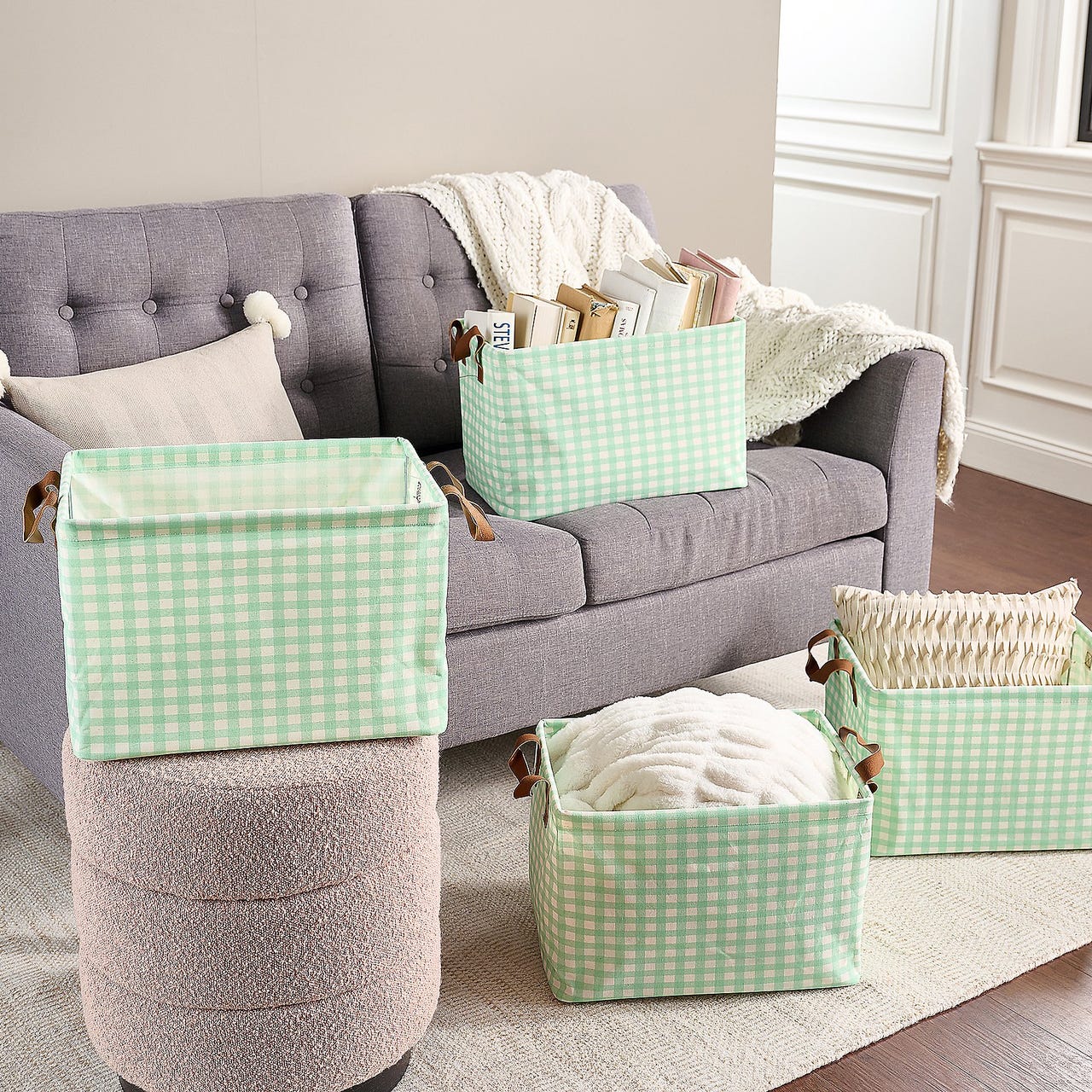 4-piece collapsible storage basket set with light green and white gingham pattern, featuring brown leather-like handles, used for organizing items like books, pillows, and blankets on and around a gray sofa.