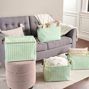 4-piece collapsible storage basket set with light green and white gingham pattern, featuring brown leather-like handles, used for organizing items like books, pillows, and blankets on and around a gray sofa.