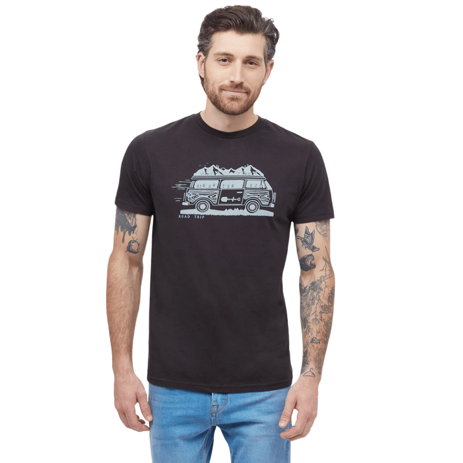 A man is wearing a black T-shirt with a \“Road Trip\“ graphic featuring a van and mountains, paired with light blue jeans. His arms show various tattoos.