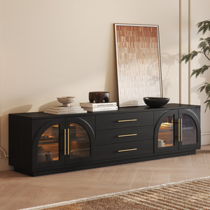 A sleek black sideboard with gold handles and arched glass doors, featuring three central drawers. Decorated with stacked books, bowls, and a leaning framed artwork.