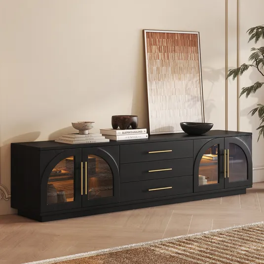 A sleek black sideboard with gold handles and arched glass doors, featuring three central drawers. Decorated with stacked books, bowls, and a leaning framed artwork.