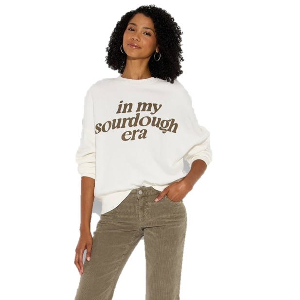 Woman wearing a white sweatshirt with the text \“in my sourdough era\“ and olive-green corduroy pants, accessorized with hoop earrings.