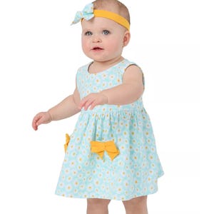 A baby wearing a sleeveless light blue dress with a daisy pattern and yellow bows, paired with a matching headband.