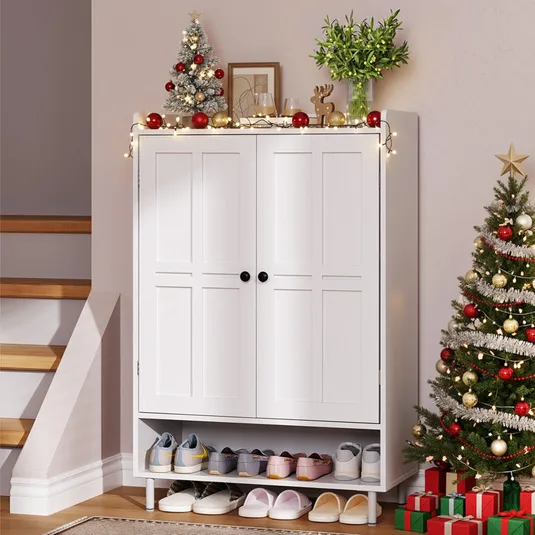 A white cabinet with shoe storage underneath is adorned with holiday decorations, including small Christmas trees, string lights, and festive ornaments. Nearby, a larger Christmas tree stands beside a stack of wrapped gifts.