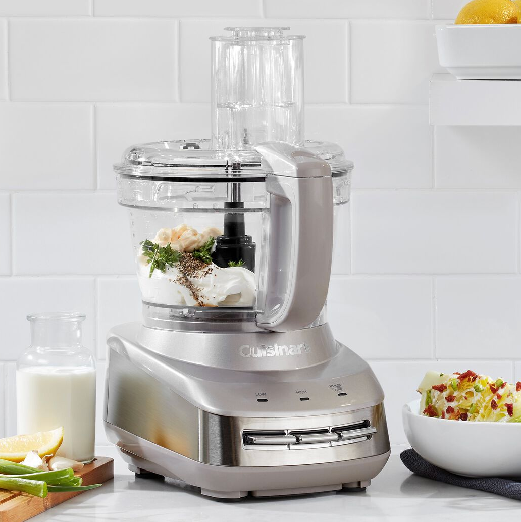 A Cuisinart food processor with ingredients inside is on a kitchen counter, surrounded by a small bottle of milk, lemon slices, green onions, and a bowl of salad.