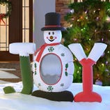 Inflatable decoration shaped like a snowman with a top hat and the letters \“JOY\“ on either side, set in a snowy holiday scene with Christmas lights.