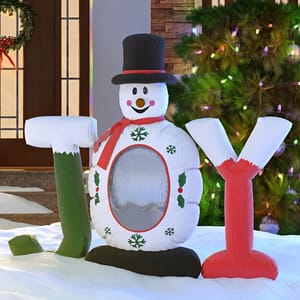Inflatable decoration shaped like a snowman with a top hat and the letters \“JOY\“ on either side, set in a snowy holiday scene with Christmas lights.
