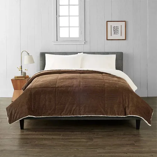 A bed with a gray upholstered headboard, white sheets, and a brown quilted comforter is in a room with white paneled walls, a wooden nightstand, a lamp, and a framed picture.