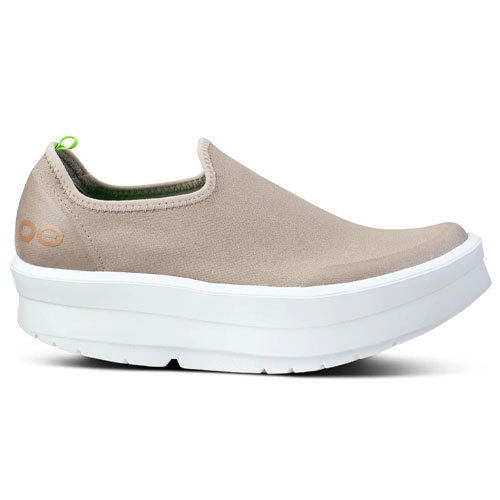 A beige slip-on shoe with a thick, white sole and a green pull tab on the heel featuring a minimalist design.