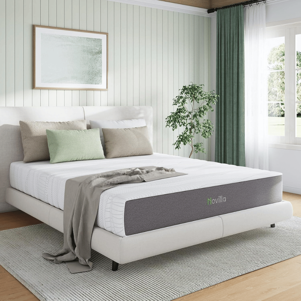 Bedroom with a white and gray bed featuring a Novilla mattress, light-colored bedding, pastel pillows, a gray throw, and a green plant. The room has a soft color palette and natural light from a window with dark green curtains.