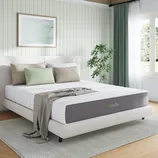 Bedroom with a white and gray bed featuring a Novilla mattress, light-colored bedding, pastel pillows, a gray throw, and a green plant. The room has a soft color palette and natural light from a window with dark green curtains.