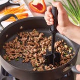 A hand holds a meat masher, breaking up ground meat in a nonstick pan on a stove.