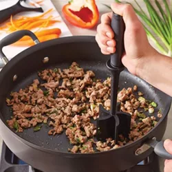A hand holds a meat masher, breaking up ground meat in a nonstick pan on a stove.