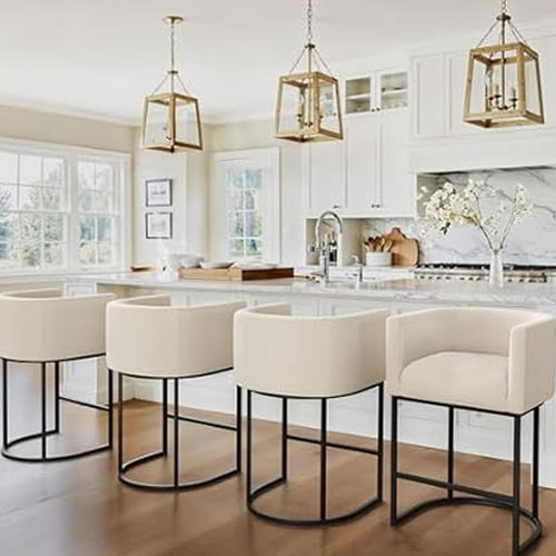 A modern kitchen features four cream-colored upholstered bar stools with metal frames, an island countertop, pendant lighting, and white cabinetry.
