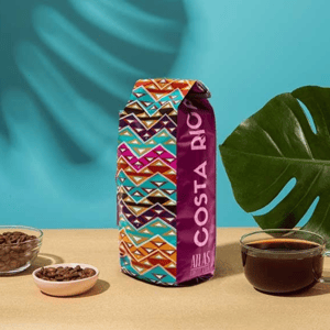A vibrantly patterned bag labeled \“COSTA RICA ATLAS COFFEE\“ beside a clear cup of coffee and two small bowls of coffee beans.
