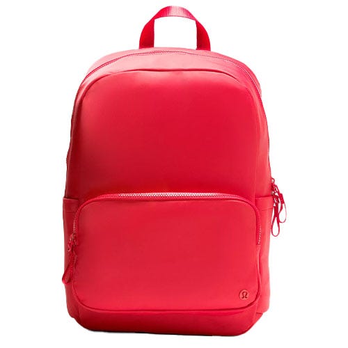 A bright red backpack with a front zippered pocket, side pocket, and a top handle, featuring a small circular logo at the bottom corner.