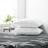 Two white pillows on a bed with gray sheets, next to a small round table holding plants and a jar, with a window in the background.