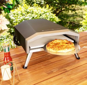A countertop outdoor pizza oven in gray and silver, with a partially cooked pizza inside, sits on a wooden table beside a bottle of sauce and a candle holder.