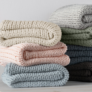 Stack of neatly folded, chunky-knit blankets in various colors, including cream, pink, blue, green, and brown, displayed against a plain background.