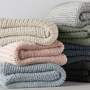 Stack of neatly folded, chunky-knit blankets in various colors, including cream, pink, blue, green, and brown, displayed against a plain background.