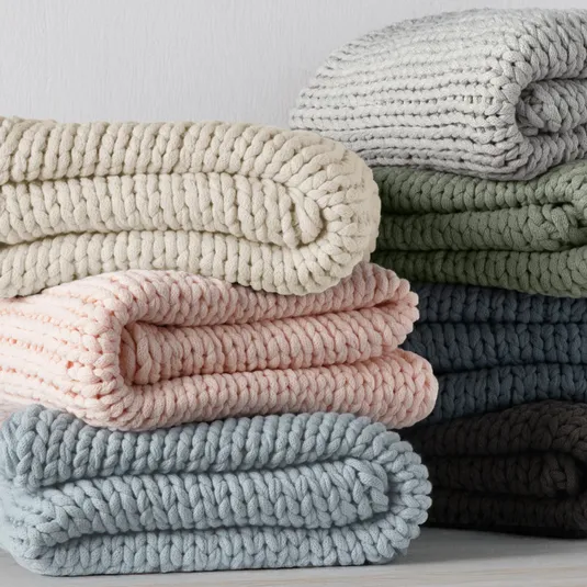Stack of neatly folded, chunky-knit blankets in various colors, including cream, pink, blue, green, and brown, displayed against a plain background.