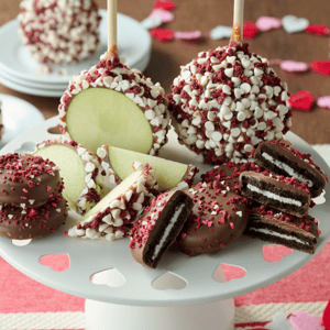 Caramel apples covered in white chocolate chips and red sprinkles sit alongside chocolate-covered sandwich cookies with similar decorations on a white plate with heart cutouts.