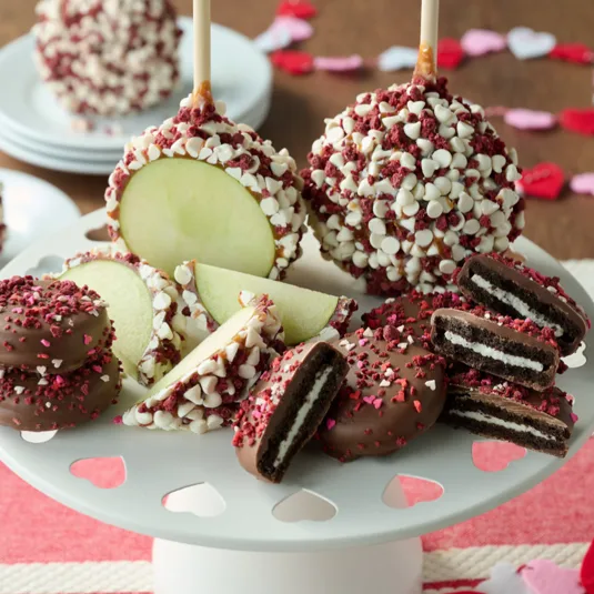 Caramel apples covered in white chocolate chips and red sprinkles sit alongside chocolate-covered sandwich cookies with similar decorations on a white plate with heart cutouts.