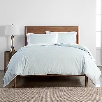 3-piece queen duvet set in light blue includes a duvet cover and two matching pillow shams, styled on a wooden bed with a white lamp beside it, placed on a woven rug.
