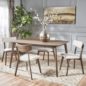A modern dining set featuring a wooden table with four chairs, all with light upholstery. The setting includes a large decorative vase with white flowers, a green plant, and a textured area rug.