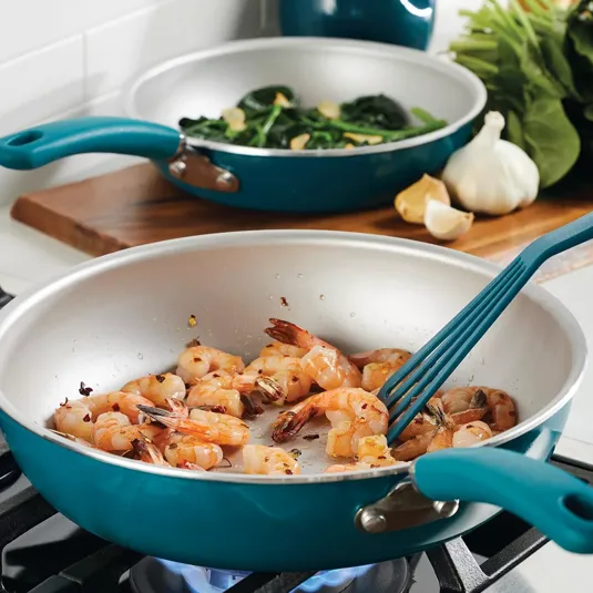Shrimp is being sautéed in a blue frying pan on a gas stove, with sautéed greens in another pan in the background alongside garlic and leafy vegetables.