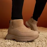 Tan ankle boots with a modern design, featuring a textured sole and smooth fabric, worn on a sandy surface with ribbed black pants.