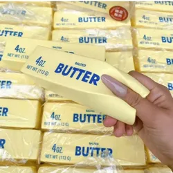 A hand holds a squishy toy resembling a stick of salted butter, labeled \“4 oz, Net Wt. 113 g,\“ among stacks of similar packages.