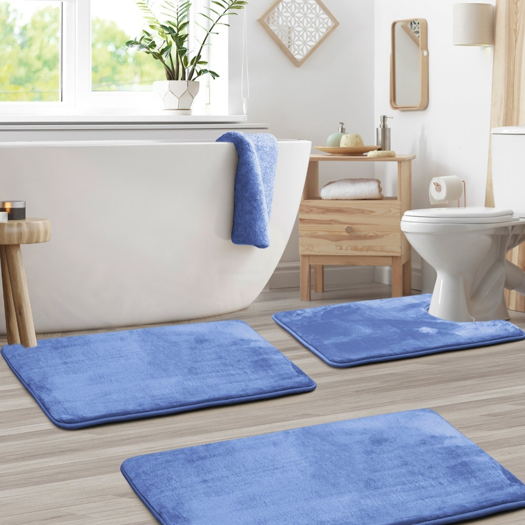 Blue bath mats are placed around a white bathtub and toilet in a modern bathroom with light wood accents, including a small wooden cabinet and stool. A potted plant sits on the windowsill.