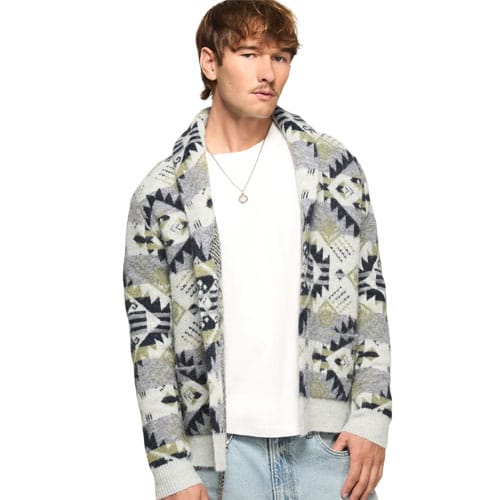 A man wearing a patterned knit cardigan with a geometric design and a white T-shirt, paired with light blue jeans, accessorized with a simple necklace.