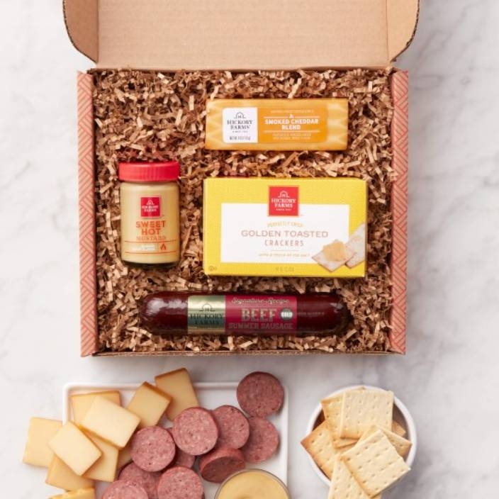 The Hickory Farms Snack Sampler includes smoked cheddar blend, golden toasted crackers, sweet hot mustard, and beef summer sausage, all arranged in a gift box with shredded packaging material.