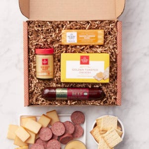 The Hickory Farms Snack Sampler includes smoked cheddar blend, golden toasted crackers, sweet hot mustard, and beef summer sausage, all arranged in a gift box with shredded packaging material.