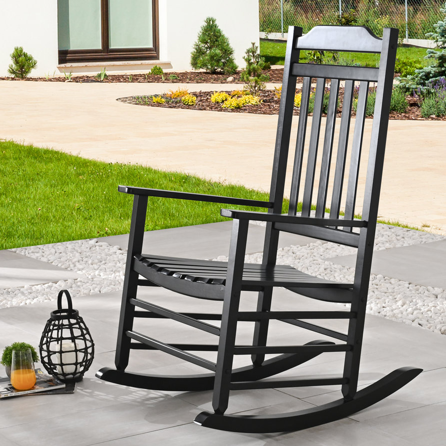A black wooden rocking chair is positioned on an outdoor patio, surrounded by greenery and decorative pebbles. Nearby are a lantern candle holder and a glass of orange juice on magazines.