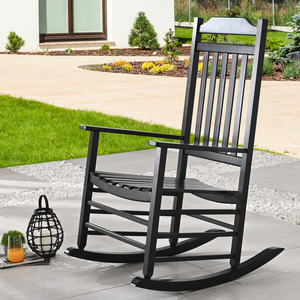 A black wooden rocking chair is positioned on an outdoor patio, surrounded by greenery and decorative pebbles. Nearby are a lantern candle holder and a glass of orange juice on magazines.