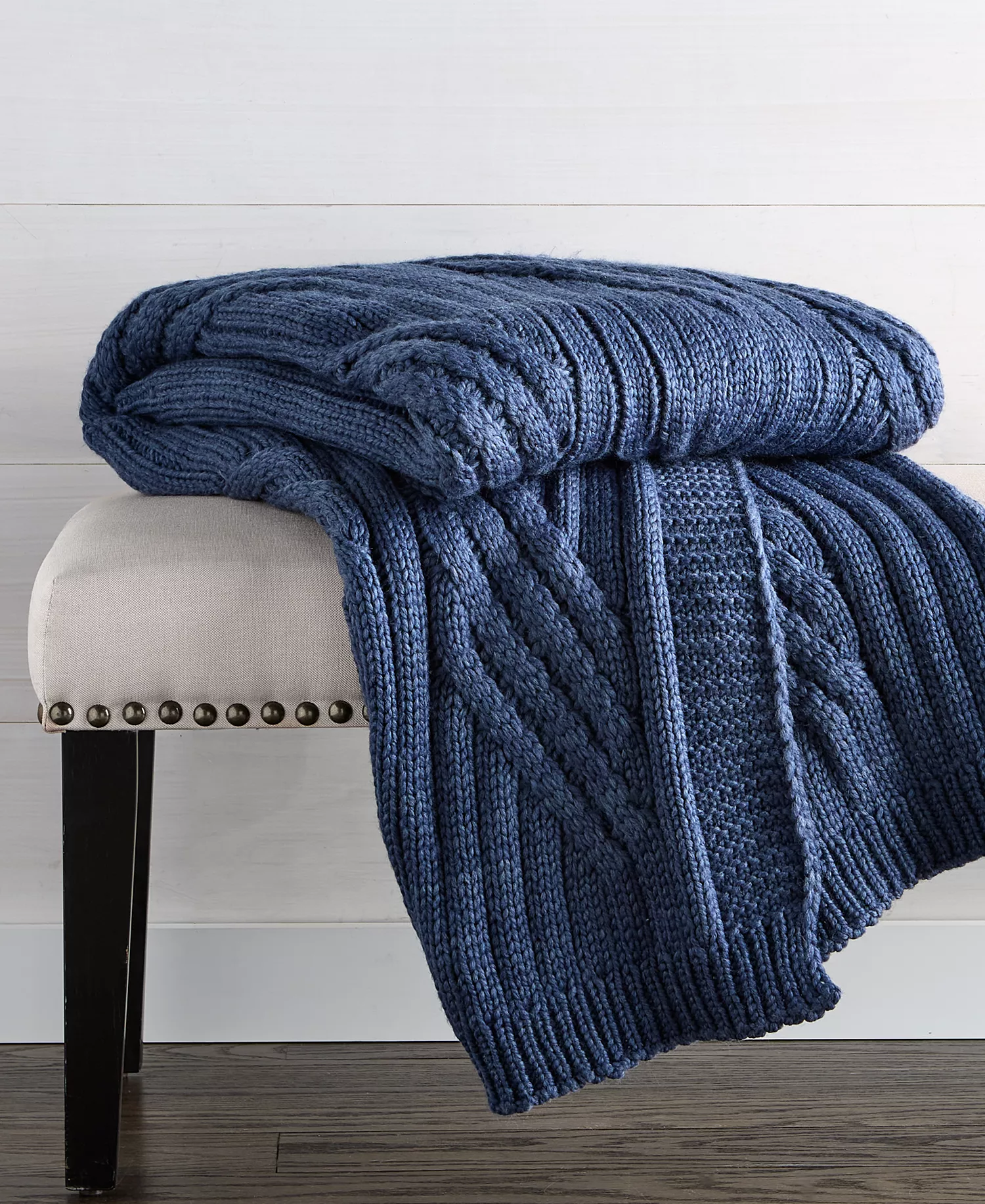 A blue cable-knit throw blanket is draped over a beige upholstered bench with dark wooden legs and nailhead trim, set on a wooden floor with a light-colored wall backdrop.