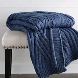 A blue cable-knit throw blanket is draped over a beige upholstered bench with dark wooden legs and nailhead trim, set on a wooden floor with a light-colored wall backdrop.