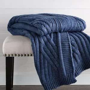 A blue cable-knit throw blanket is draped over a beige upholstered bench with dark wooden legs and nailhead trim, set on a wooden floor with a light-colored wall backdrop.
