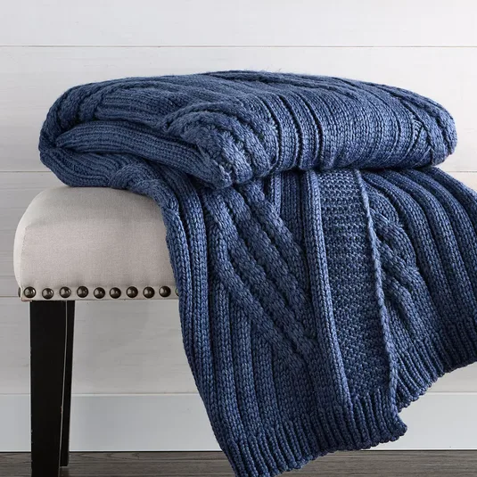 A blue cable-knit throw blanket is draped over a beige upholstered bench with dark wooden legs and nailhead trim, set on a wooden floor with a light-colored wall backdrop.