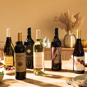 A collection of six wine bottles displayed on a wooden table, featuring a mix of red and white wines with various labels and designs, set against a neutral interior background.