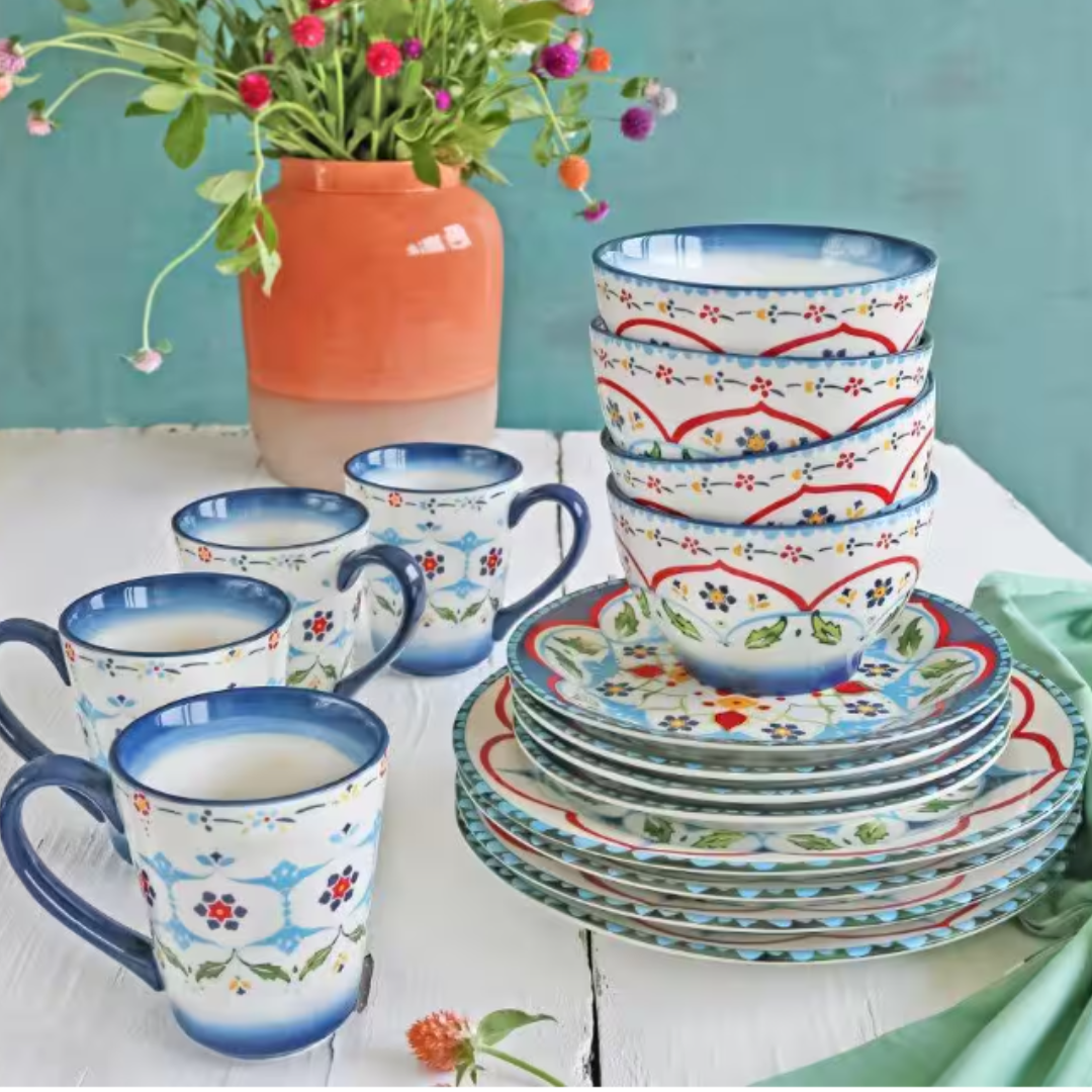 A 16-piece stoneware set with a blue and red floral pattern; includes bowls, plates, and mugs on a white table next to a terracotta vase with flowers.