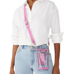 A model is wearing a clear phone crossbody bag with pink leather trim and an adjustable strap, paired with a white shirt and light blue jeans.