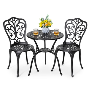 A 3-piece patio bistro set featuring two black, ornate metal chairs and a matching round table with a decorative design. The table has a small vase with yellow flowers and two glasses on top.