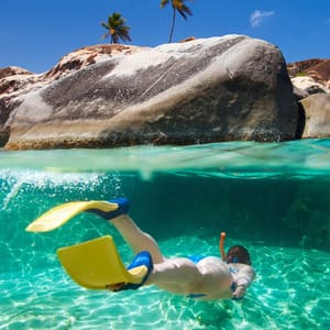 A person is snorkeling in clear turquoise water, wearing a snorkel, goggles, and yellow fins, near large rocks and palm trees.